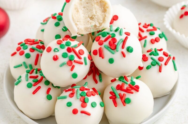 A plate of sugar cookie truffles, each topped with festive red and green sprinkles. One truffle is bitten, revealing a creamy interior. White paper cups and a bowl of sprinkles are in the background.